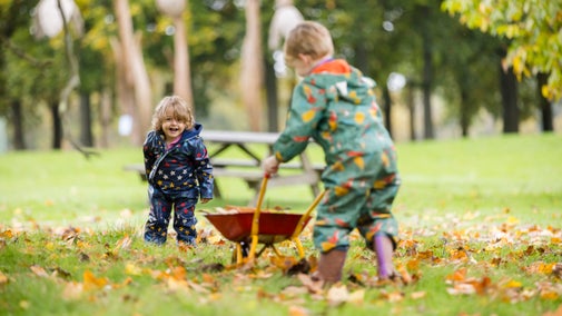 Two children in colourful romper suits collect autumn leaves and put them in a mini wheelbarrow